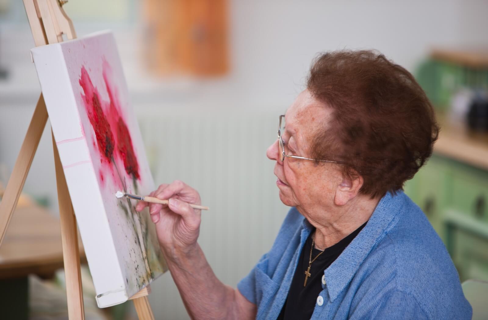 An older adult woman painting a flower on a canvas.
