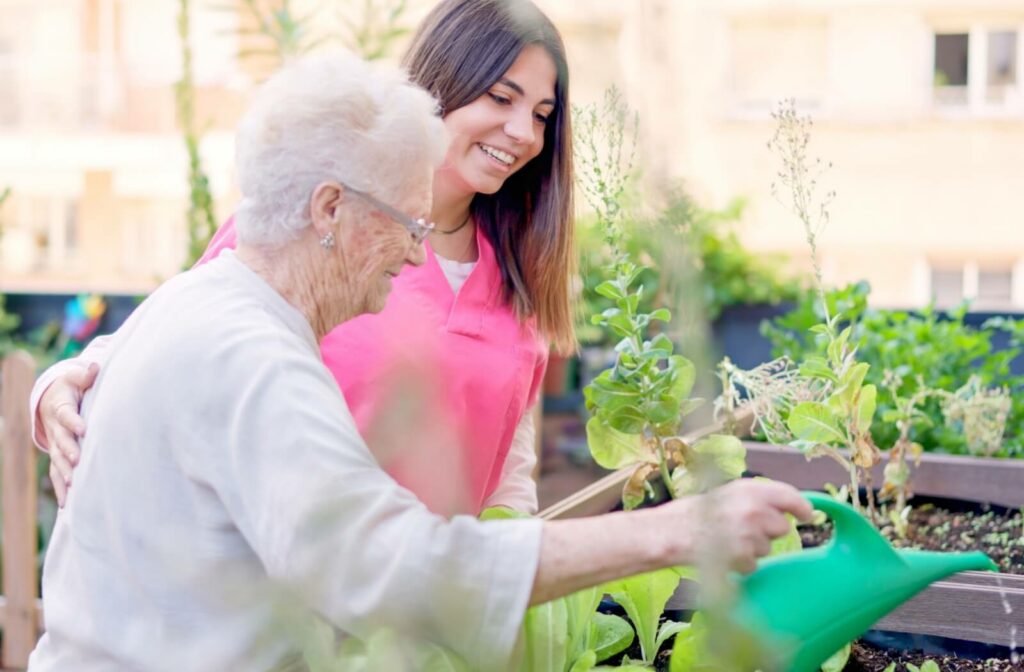 A memory care staff member assists an older adult water the community garden.
