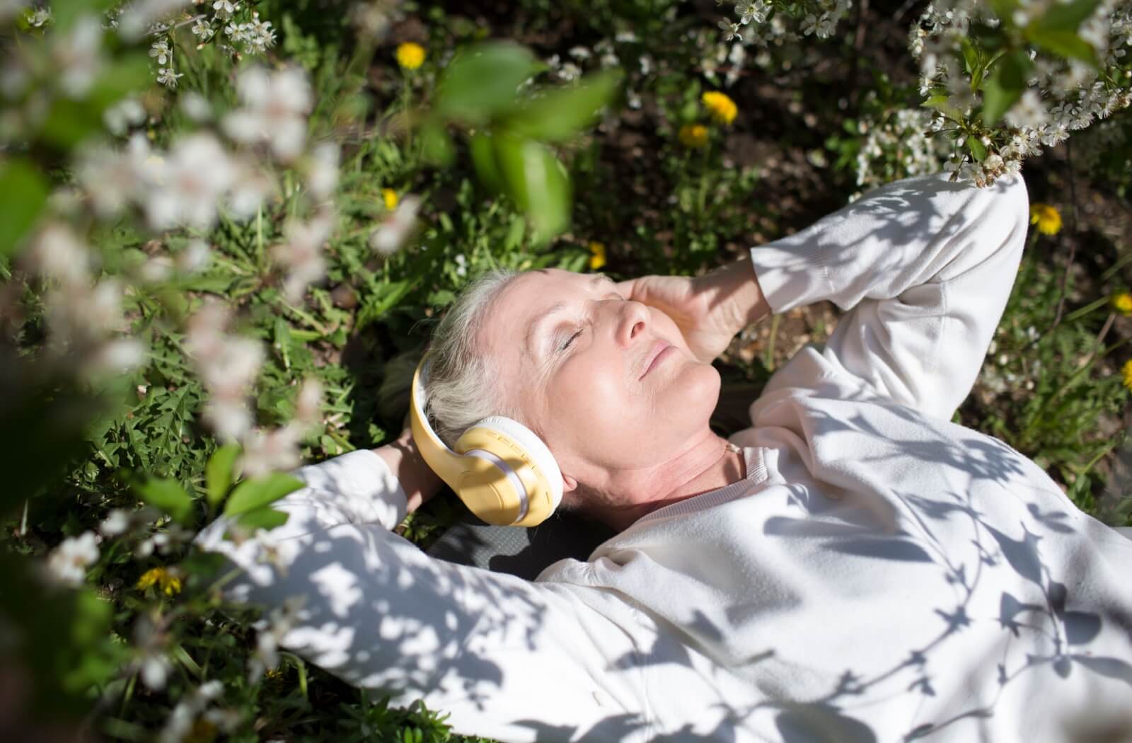 An older adult wearing yellow headphones lies contentedly in the grass with the sun shining warm light on their face.