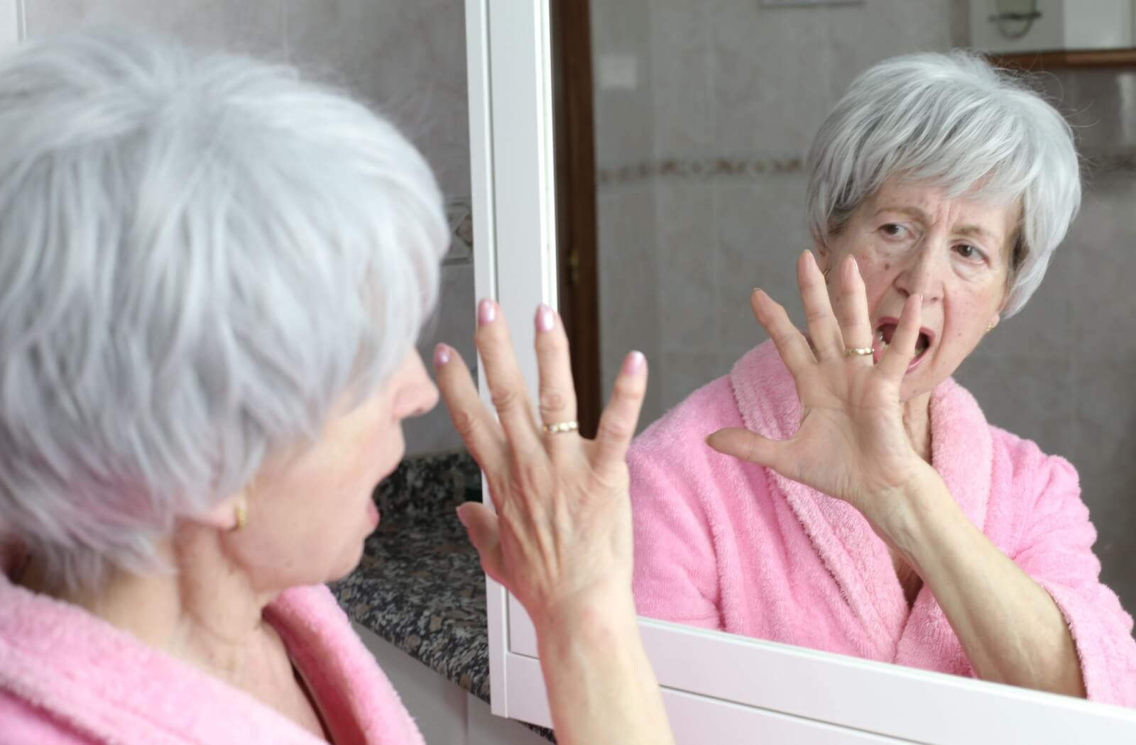 A mature woman raises her hand to her face, startled by her own reflection as a hallucination takes her by surprise.