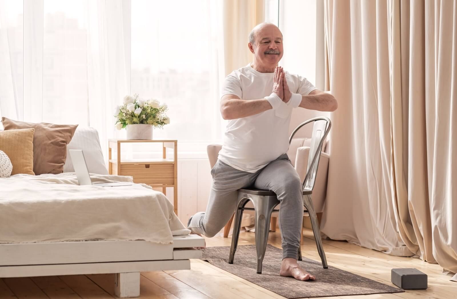 An older adult uses a chair to support themself while performing a lunging pose while doing yoga in their home.