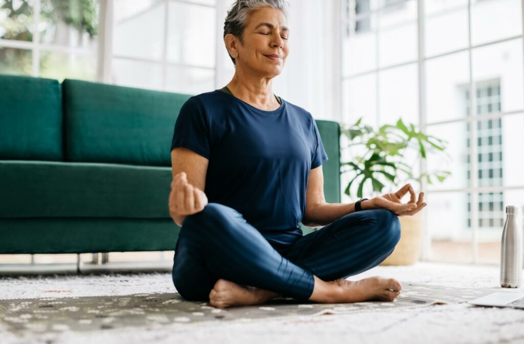 A peaceful older adult meditates on the floor of their living room after yoga to promote mindfulness and reduce stress.