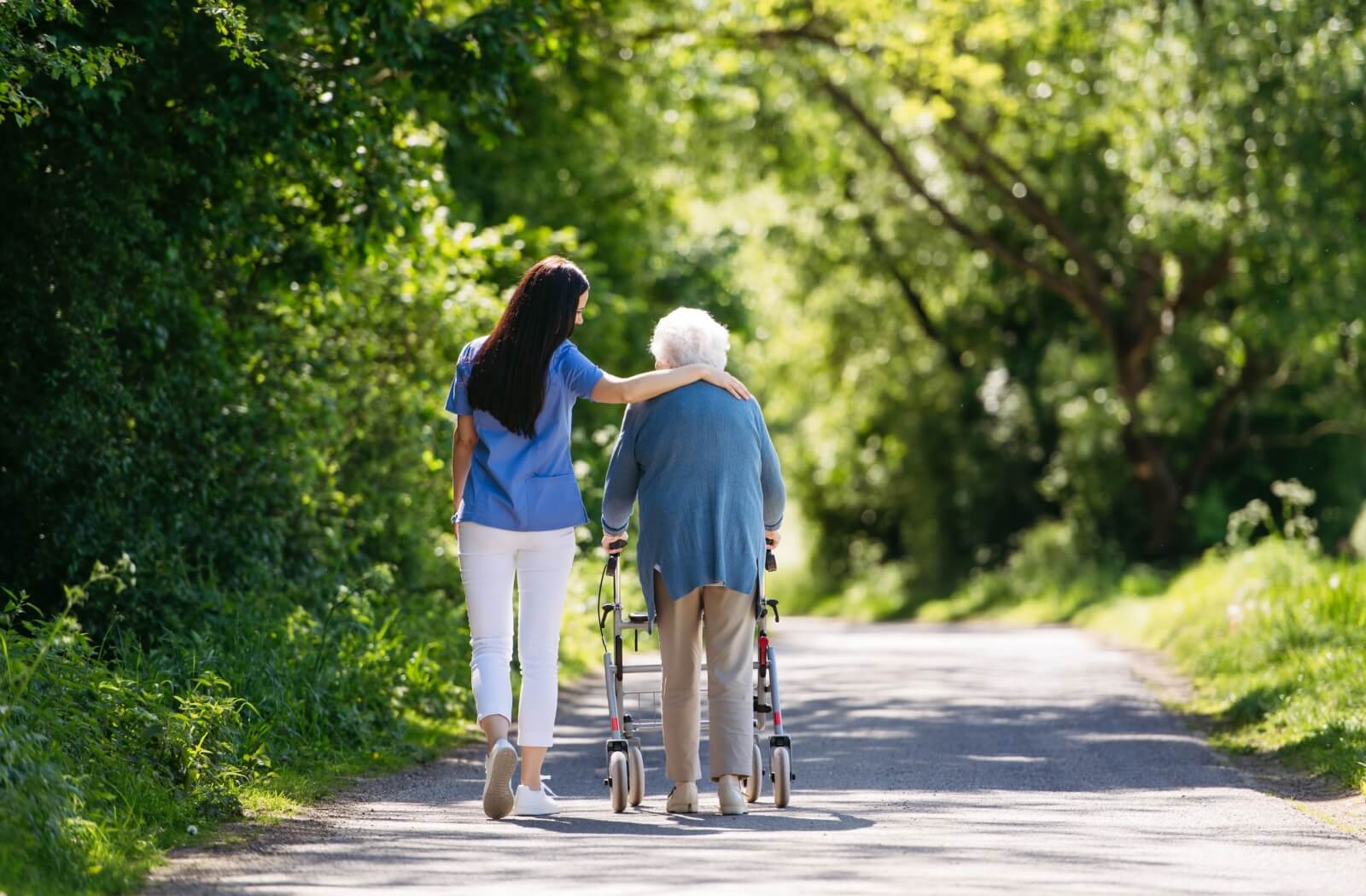A shot from behind of a caregiver and an older adult in senior living walking together on a nature path.