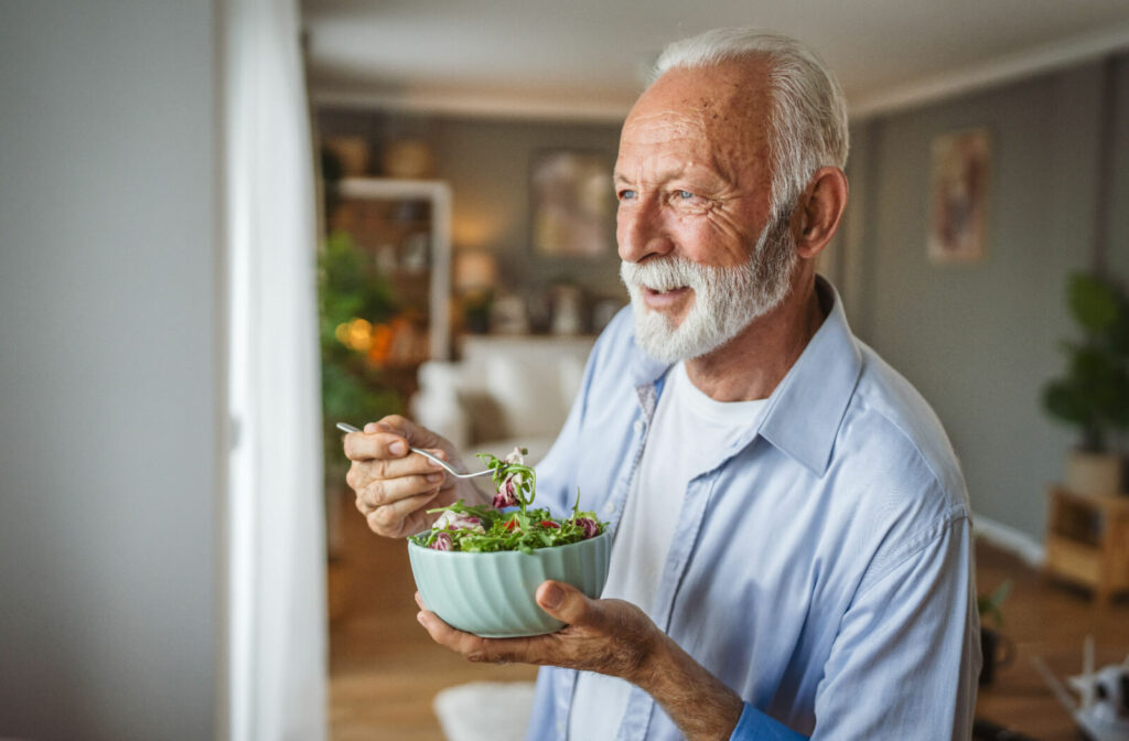 An older adult smiles while taking a bite of salad and looking out the window of their home in senior living