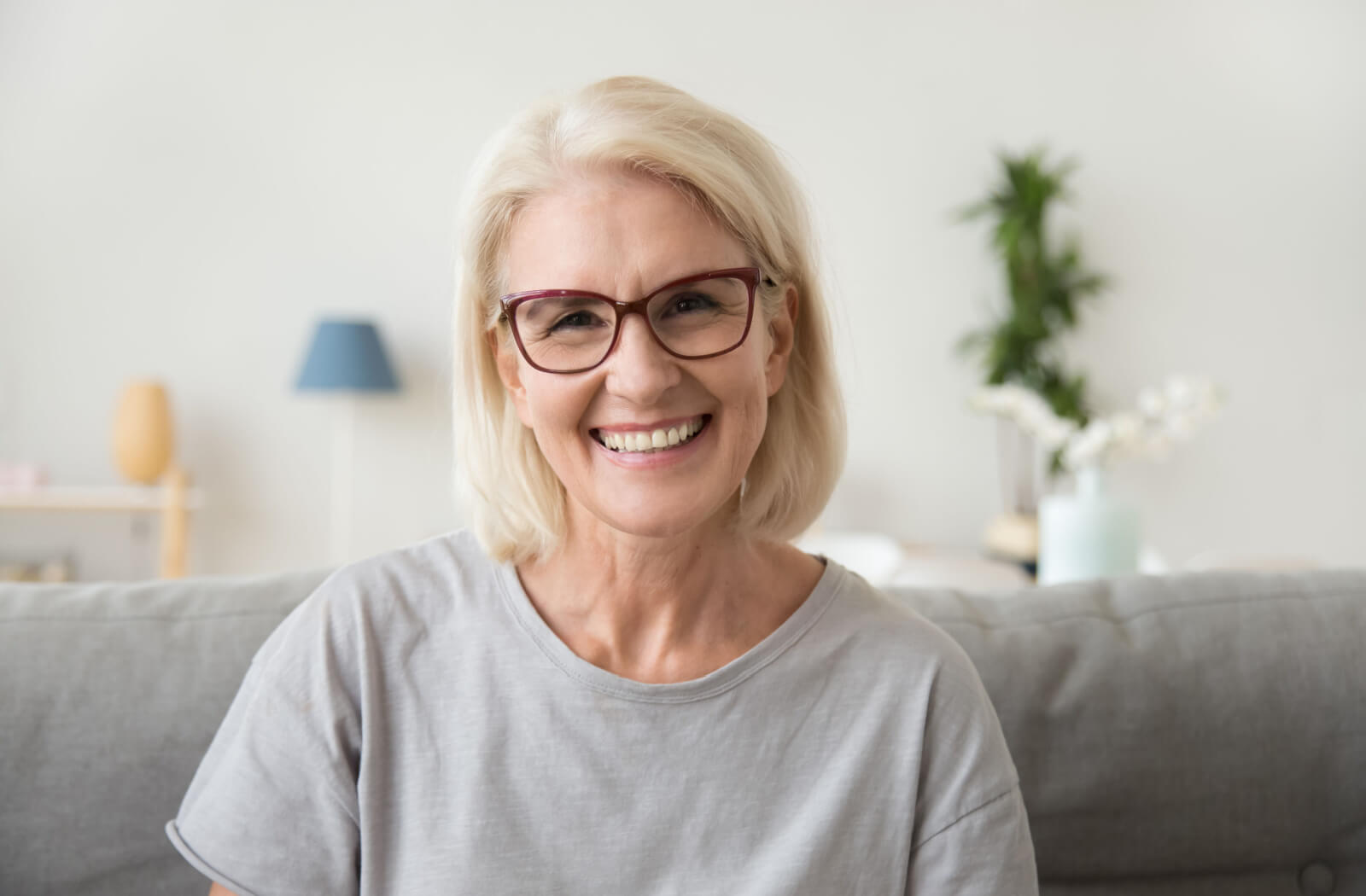 An older adult woman with glasses, sitting on the couch, smiling and looking directly at the camera