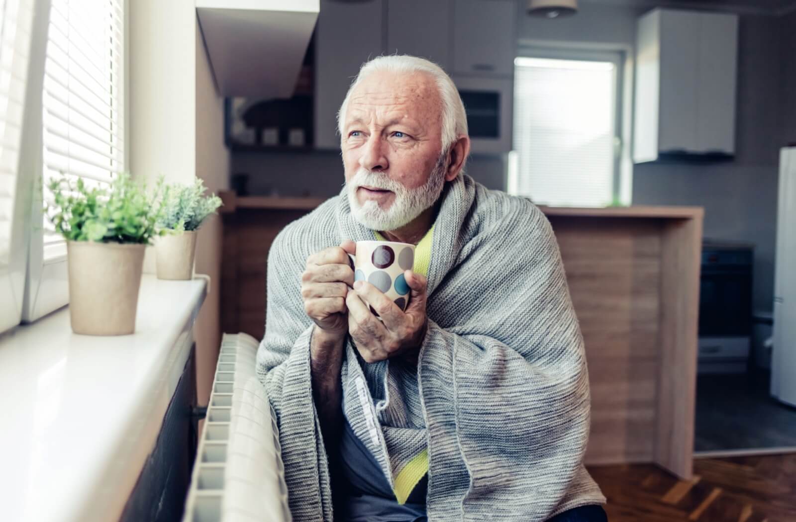 An older adult holds a warm mug while covered in a blanket, sitting next to the radiator, in an effort to keep warm