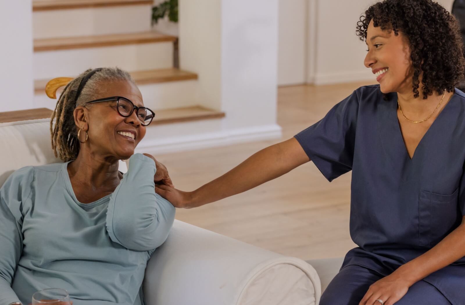 Smiling older adult sitting on sofa with care team member gently touching shoulder in supportive conversation