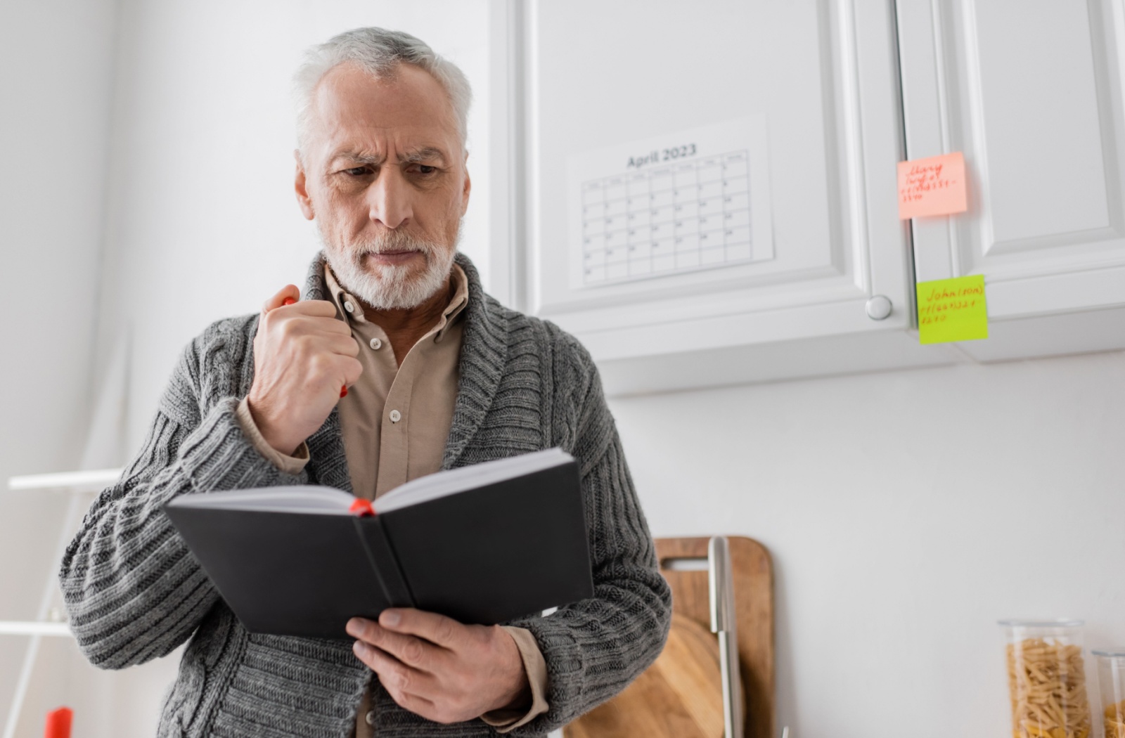 An older adult in a kitchen stares in confusion at a day planner in front of a calendar with reminder sticky notes attached.