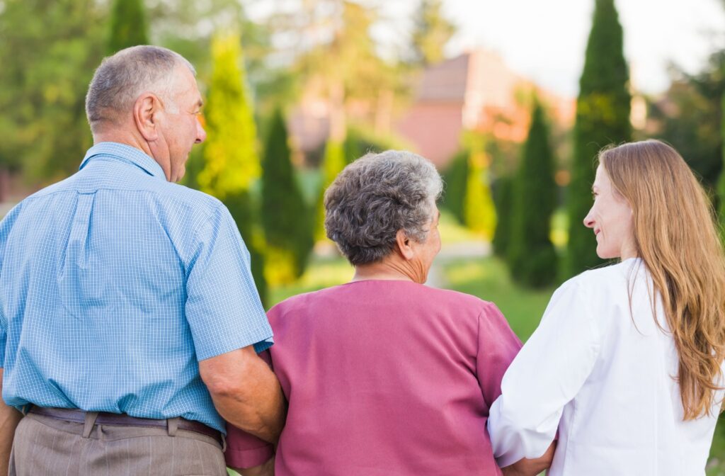 An older adult couple walks arm-in-arm with a memory care worker outdoors, connecting with nature as part of their therapy.