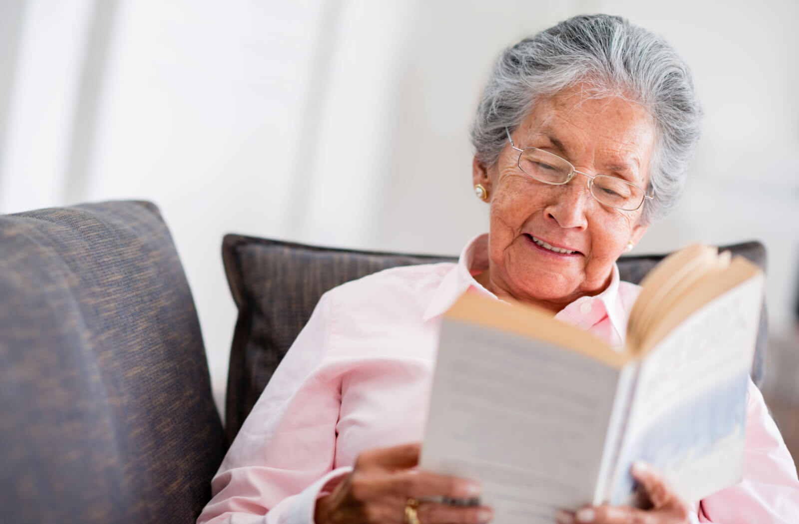 A happy older adult woman reading a book while sitting on a couch.