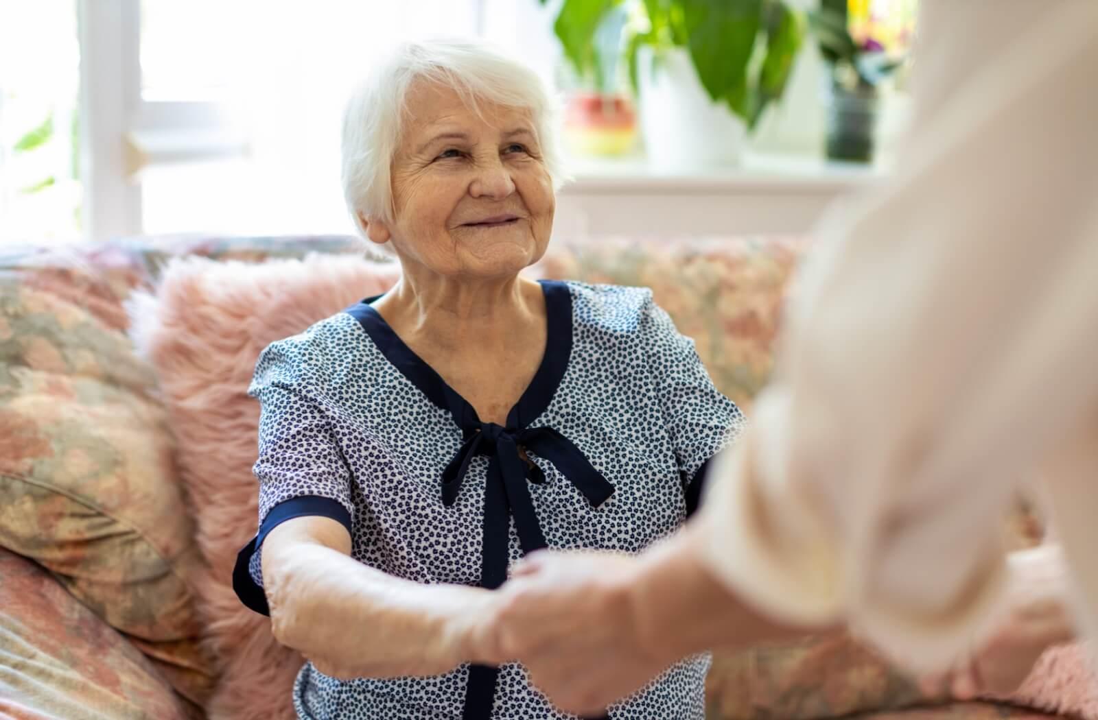 A caregiver (off camera) helps a senior stand up from a sitting position.